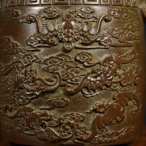Puede incluir: Primer plano de un jarr&oacute;n de bronce con intrincados tallados de dragones y nubes. El jarr&oacute;n tiene una p&aacute;tina marr&oacute;n oscura y una superficie texturizada.