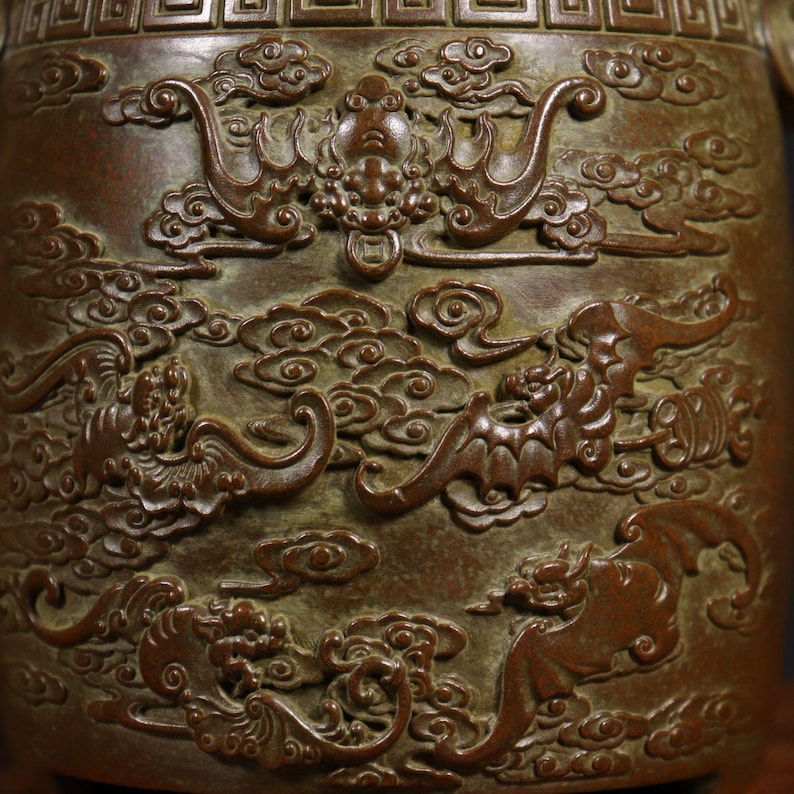 Puede incluir: Primer plano de un jarr&oacute;n de bronce con intrincados tallados de dragones y nubes. El jarr&oacute;n tiene una p&aacute;tina marr&oacute;n oscura y una superficie texturizada.