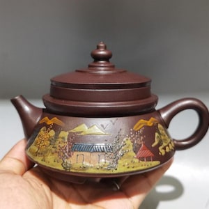 以下が含まれることがあります： 蓋と取っ手が付いた茶色のセラミックティーポット。 ティーポットは、黄色、茶色、青の色合いの木、家、鳥を特徴とする風景シーンで装飾されています。 ティーポットは手で持たれています。