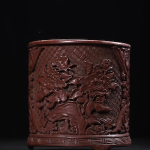 May include: A cylindrical, reddish-brown container with intricate floral carvings. The detailed design features large peony blooms and foliage set against a crosshatched background. The container has a decorative base and is likely a decorative item.