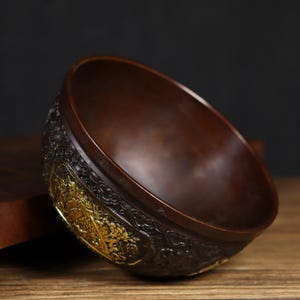 May include: A dark brown bowl with intricate gold detailing. The bowl is made of metal and has a rounded shape. The bowl is sitting on a wooden surface.