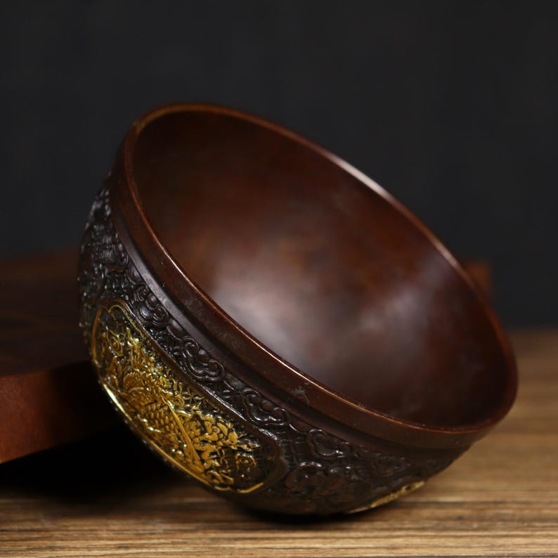 May include: A dark brown bowl with intricate gold detailing. The bowl is made of metal and has a rounded shape. The bowl is sitting on a wooden surface.