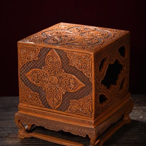 May include: A square wooden box with intricate relief carvings featuring floral motifs and geometric patterns. The box has a textured brown surface, a removable lid with a central carved design, and sits on a decorative carved wooden base.