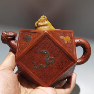 以下が含まれることがあります： 猿の形をした蓋が付いた、赤茶色の幾何学的なティーポット。 ティーポットには動物のイラストと竜の形をした注ぎ口があります。 ハンドルは円形です。 ティーポットは手で持たれています。