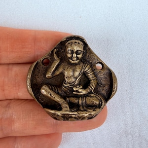 May include: A bronze-colored metal Buddha pendant. The pendant features a seated Buddha figure in a meditative pose, with intricate details. The pendant has two holes for hanging and is set against a white background.