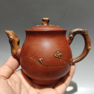 May include: A small, reddish-brown ceramic teapot with a wooden handle and spout. The teapot features a lid with a wooden knob and floral designs. The handle and spout are made to look like tree branches.