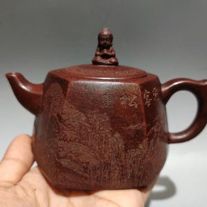 May include: A dark brown hexagonal clay teapot featuring an engraved landscape scene on the side, Chinese characters, and a small sculpted figure of a seated person with hands pressed together in a prayer position serving as the lid handle.