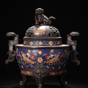 Op de afbeelding: Een sierlijke, antieke wierookbrander met een donkerblauwe emaille basis, versierd met bloemen- en drakenmotieven in rood, geel en wit. Een kleine, bronskleurige leeuwenfiguur zit bovenop het geperforeerde deksel. De brander heeft drie poten.