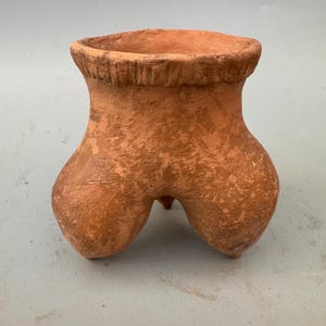 May include: A terracotta pot with three legs, shaped like a stylized human figure. The pot has a rough, textured surface and a slightly flared rim.