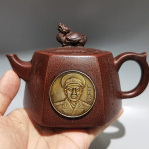 May include: A hexagonal brown clay teapot featuring a circular metallic medallion on the front depicting a military figure in uniform with the text 徐向前元帅. The lid is topped with a small sculpted creature, and the sides are engraved with Chinese characters.