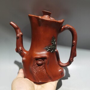 Può includere: Una teiera marrone-rossastra con un design a tronco d'albero. Il beccuccio e il manico imitano i rami e un motivo di foglie scure è sul lato. La teiera ha un coperchio e una superficie strutturata, creando un'estetica rustica.