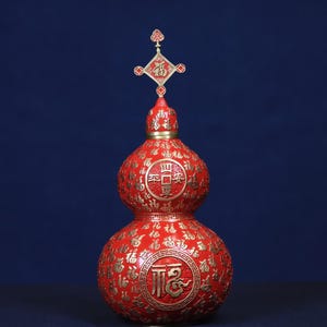 May include: A red gourd-shaped vessel featuring gold-colored Chinese characters and decorative patterns. The surface is covered in repeating symbols, with a prominent circular emblem on the body and a metal finial topper featuring the character Fu.