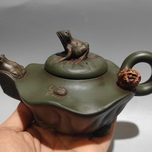 Puede incluir: Una tetera artesanal verde oliva con temática de rana. La tetera tiene forma de hoja de nenúfar, con una rana en la tapa y otra en el pico. El asa tiene un adorno de madera tallada. Una pieza única para los amantes del té.