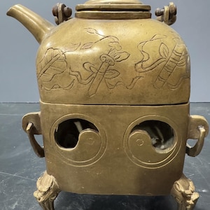 May include: A brass teapot with a decorative design and a stand with three legs. The teapot has a lid and a spout. The stand has two circular openings on the sides.