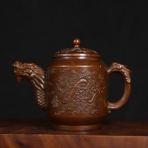 Könnte beinhalten: Eine kleine, braune, kupferne Teekanne mit einem Drachenmuster. Die Teekanne hat einen Deckel mit einem Knopf oben und einem drachenförmigen Ausguss. Die Teekanne ist mit einem Drachen und Wolken verziert.
