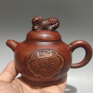 May include: A brown clay teapot featuring a rounded body, a curved handle, and a short spout. The lid is topped with a sculpted animal figure. A central decorative plaque on the front displays embossed Chinese characters in a rectangular frame.