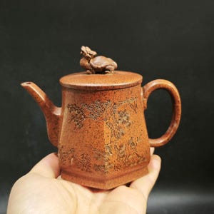 Könnte beinhalten: Braune Keramik-Teekanne mit Deckel und Henkel. Die Teekanne hat detaillierte Schnitzereien einer Landschaft mit Bäumen und Figuren. Eine kleine Schildkrötenfigur ziert den Deckel. Die Teekanne wird in einer Hand gehalten.