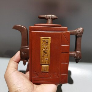 May include: A reddish-brown, rectangular teapot with a curved spout and a handle. The teapot has a decorative handle and a lid with a small knob. A rectangular gold-colored plaque with text is on the front.