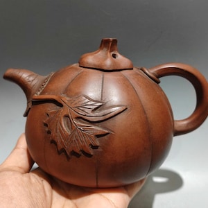 以下が含まれることがあります： 垂直の隆起があるカボチャの形をした茶色の粘土製ティーポット。湾曲したハンドル、短い注ぎ口、茎のようなつまみが付いた蓋が特徴です。丸みを帯びた本体の側面には、彫り込まれた葉のデザインがはっきりと施されています。