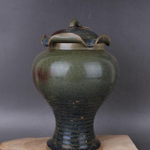 May include: A green and blue ceramic jar with a lid. The jar has a wide base and a narrow neck. The lid has a scalloped edge. The jar is sitting on a wooden surface.