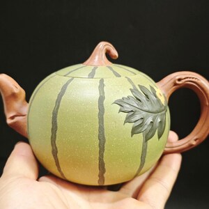 May include: A ceramic teapot in the shape of a watermelon, held in a hand. The teapot is primarily light green with dark green stripes and a brown handle, spout, and lid. A leaf design is on the side.