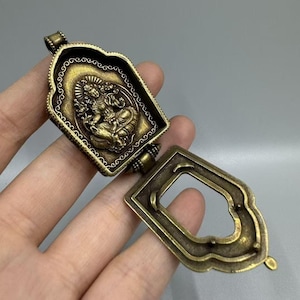 Pode incluir: Um pingente de latão antigo com uma moldura retangular aberta e uma tampa articulada. A tampa apresenta um relevo detalhado de uma divindade sentada dentro de uma borda ornamentada. O pingente é mantido aberto, revelando seu interior.