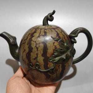 May include: A round teapot shaped like a watermelon with a dark green rind pattern, a dark brown spout, a curved handle resembling a vine, and a lid topped with a curled stem. A sculpted green leaf and vine detail are attached to the side.