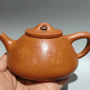 Może przedstawiać: Mały, brązowy ceramiczny imbryk z pokrywką i uchwytem. Imbryk ma zaokrąglony korpus i dzióbek do nalewania. Pokrywka ma mały otwór. Imbryk jest trzymany w dłoni.