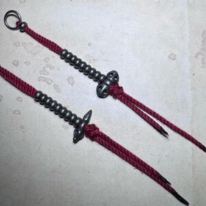 May include: Two handcrafted decorative items featuring a dark red cord, silver-coloured metal beads and accents. Each item has a loop at one end and two cord tails at the other.