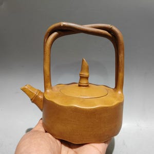 May include: A light brown ceramic teapot with a textured, scalloped body and a bamboo-inspired spout. The lid features a pointed finial, and the handle is formed by two intertwined, branch-like elements that arch over the top of the vessel.