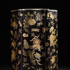 Puede incluir: Un recipiente negro octogonal con diseños florales y geométricos dorados. El recipiente presenta adornos dorados y está decorado con flores, hojas y símbolos estilizados. La estética general es ornamentada y tradicional.