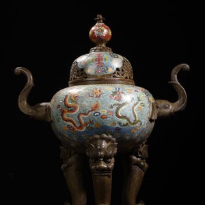 Op de afbeelding: Een antieke bronzen wierookbrander met een lichtblauwe emaille afwerking. De brander heeft een gedetailleerd drakenmotief, sierlijke handvatten en decoratieve poten. Het deksel is bekroond met een kleine, kleurrijke knop. Het algehele ontwerp is ingewikkeld en toont traditioneel vakmanschap.