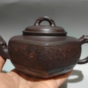 May include: A brown ceramic teapot with a lid and handle. The teapot has a decorative pattern around the body and a dragon-shaped handle. The teapot is held in a hand.