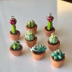 Puede incluir: Una colección de dijes de cactus en miniatura en pequeñas macetas de terracota. Los cactus son de varios tonos de verde, algunos con detalles rojos y blancos. Cada dije tiene un lazo plateado para sujetar.