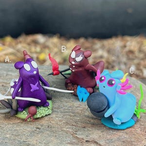 Puede incluir: Tres figuras de arcilla coloridas. Una criatura morada con ojos blancos y una estrella en el pecho, una criatura granate con un cuerno rojo y una criatura azul con detalles rosas y amarillos, cada una sobre una pequeña base.