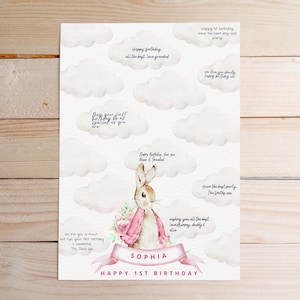 May include: A white card featuring a pink bunny rabbit illustration and clouds. The card has a pink ribbon with the name "Sophia" and the text "Happy 1st Birthday". There are blank spaces for guests to write messages.