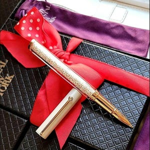 May include: A luxury pen with a gold-colored barrel and a sparkling crystal-studded upper section. The pen is presented with a red ribbon and a white polka-dotted bow, resting on a black box with a diamond pattern.