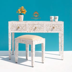 Puede incluir: Una mesa consola de nácar blanco con tres cajones y un taburete a juego con asiento tapizado en beige. La superficie de la mesa sostiene una pequeña planta amarilla en maceta y dos tazas blancas. El logotipo The Indian Furniture es visible.