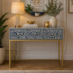 May include: A navy blue and white floral patterned console table with gold legs. The table has three drawers with gold knobs. A gold lamp, mirror, and decorative items sit on top of the table. The Indian Furniture logo is visible.