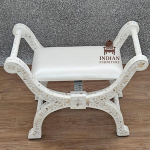 May include: A white bench with curved legs and armrests, adorned with floral and leaf patterns. The seat is a rectangular cushion. The bench is made of a light-colored material, possibly wood, and is suitable for indoor use.