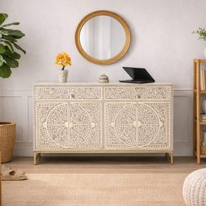 May include: A cream-colored cabinet with intricate floral patterns, gold accents, and a matching round mirror. A laptop, decorative items, and a plant are on top. A wooden bookshelf and a woven basket are also visible.