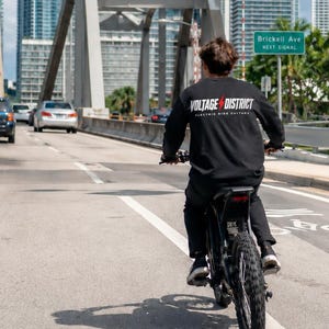 Puede incluir: Una camiseta negra de manga larga con el texto blanco VOLTAGE DISTRICT, un icono de rayo rojo y el texto más pequeño en blanco ELECTRIC RIDE CULTURE en la espalda, usada mientras se conduce una bicicleta eléctrica negra en una calle.