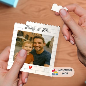 May include: A white brick-style photo frame with the words "Daddy & Me" and a photo of a man and a child. A small red heart is at the bottom. The frame is held by a person's hands, with a single white brick held in the other hand.