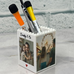 Op de afbeelding: Een witte penhouder in blokstijl met familiefoto's en de tekst "Daddy & Me". De houder bevat oranje, gele en zilveren pennen en stiften. Er staat een rood hart op de voorkant. De penhouder staat op een reflecterend oppervlak.