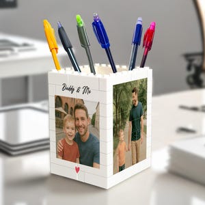 Puede incluir: Un portalápices blanco con una foto de un padre y un niño, y el texto "Daddy & Me". El portalápices contiene varios bolígrafos con tinta amarilla, negra, verde, azul y roja. Un corazón rojo está en la parte inferior de la foto.