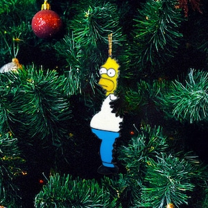 Puede incluir: Un adorno navideño de Homer Simpson. El adorno es amarillo, blanco y azul, y cuelga de un árbol de Navidad verde con adornos rojos y plateados. El adorno es una representación de dibujos animados de Homer Simpson.