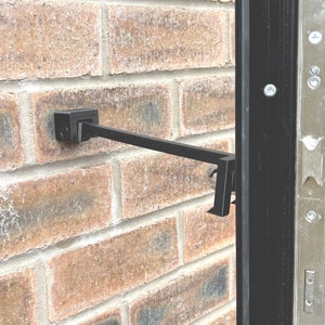 Op de afbeelding: Een zwarte metalen deurstopper bevestigd aan een bakstenen muur. De deurstopper heeft een vierkante basis en een schuine arm met een haak. De bakstenen muur is gemaakt van rode en bruine bakstenen.