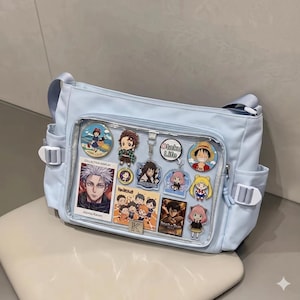 以下が含まれることがあります： アニメのキャラクターチャームとカードを表示する透明なフロントポケットが付いた、水色のメッセンジャーバッグ。バッグにはサイドポケットとショルダーストラップが付いています。チャームには、さまざまなアニメシリーズのキャラクターが含まれています。「Otaku Life」の文字が見えます。