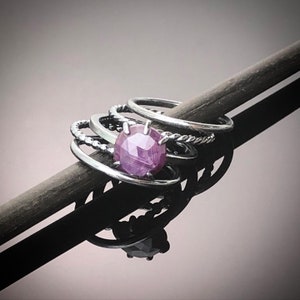 May include: A silver ring with a pink gemstone set in the center. The ring is stacked with other silver bands.