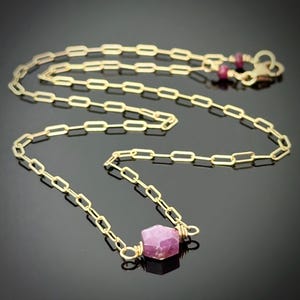 May include: A gold-colored chain necklace with a rectangular link design. The necklace features a faceted, pink gemstone pendant and two small red gemstones near the clasp. The jewelry is displayed on a dark surface.